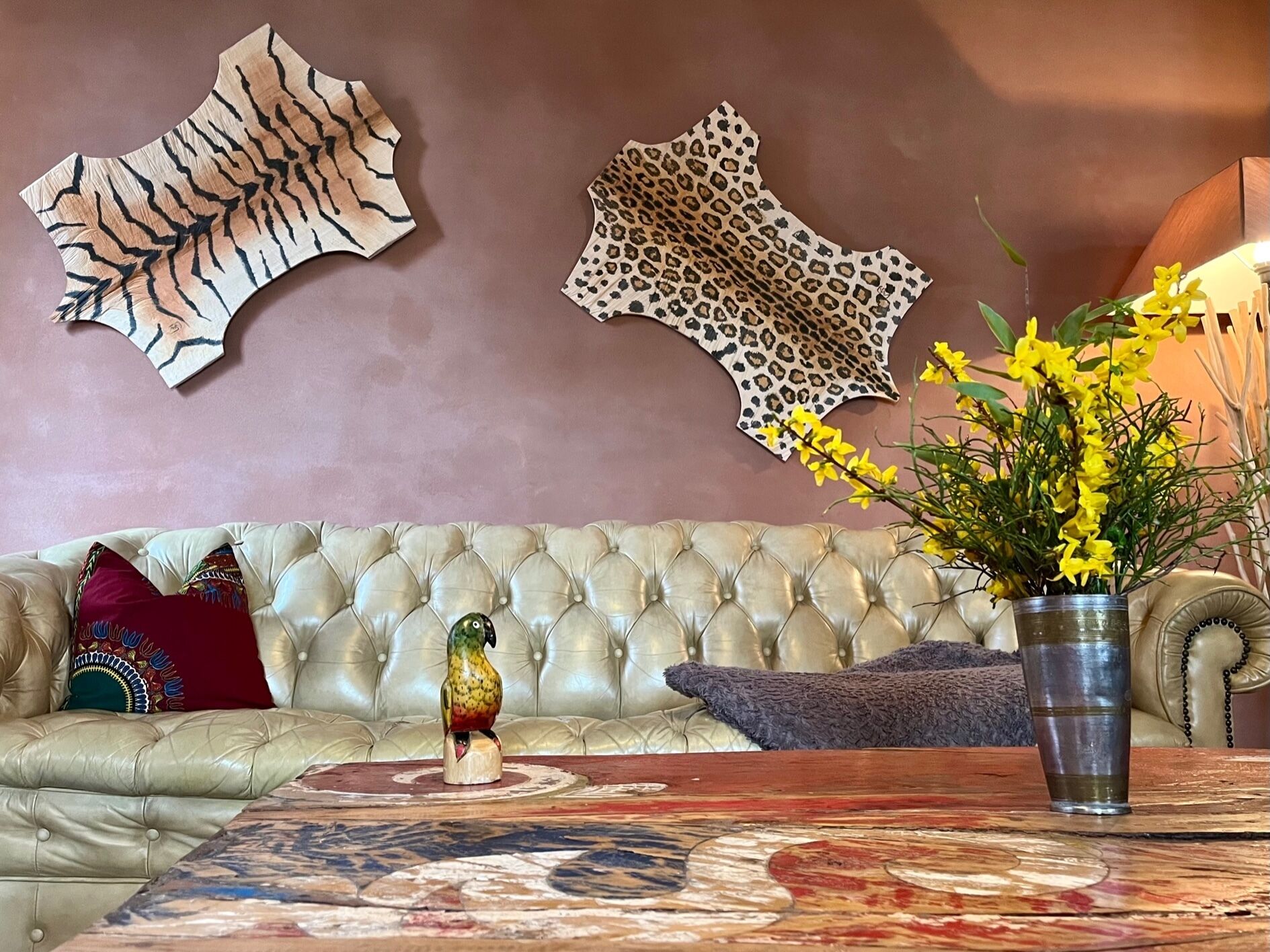 Un sofá de cuero con capitonado, almohadones decorativos, una escultura de pájaro de madera, un florero con flores amarillas y dos adornos de pared.