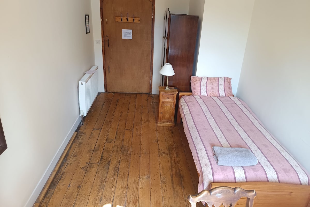 A bedroom with a wooden door, a radiator, a bedside table with a lamp, a bed with a striped bedspread, and a wooden headboard.