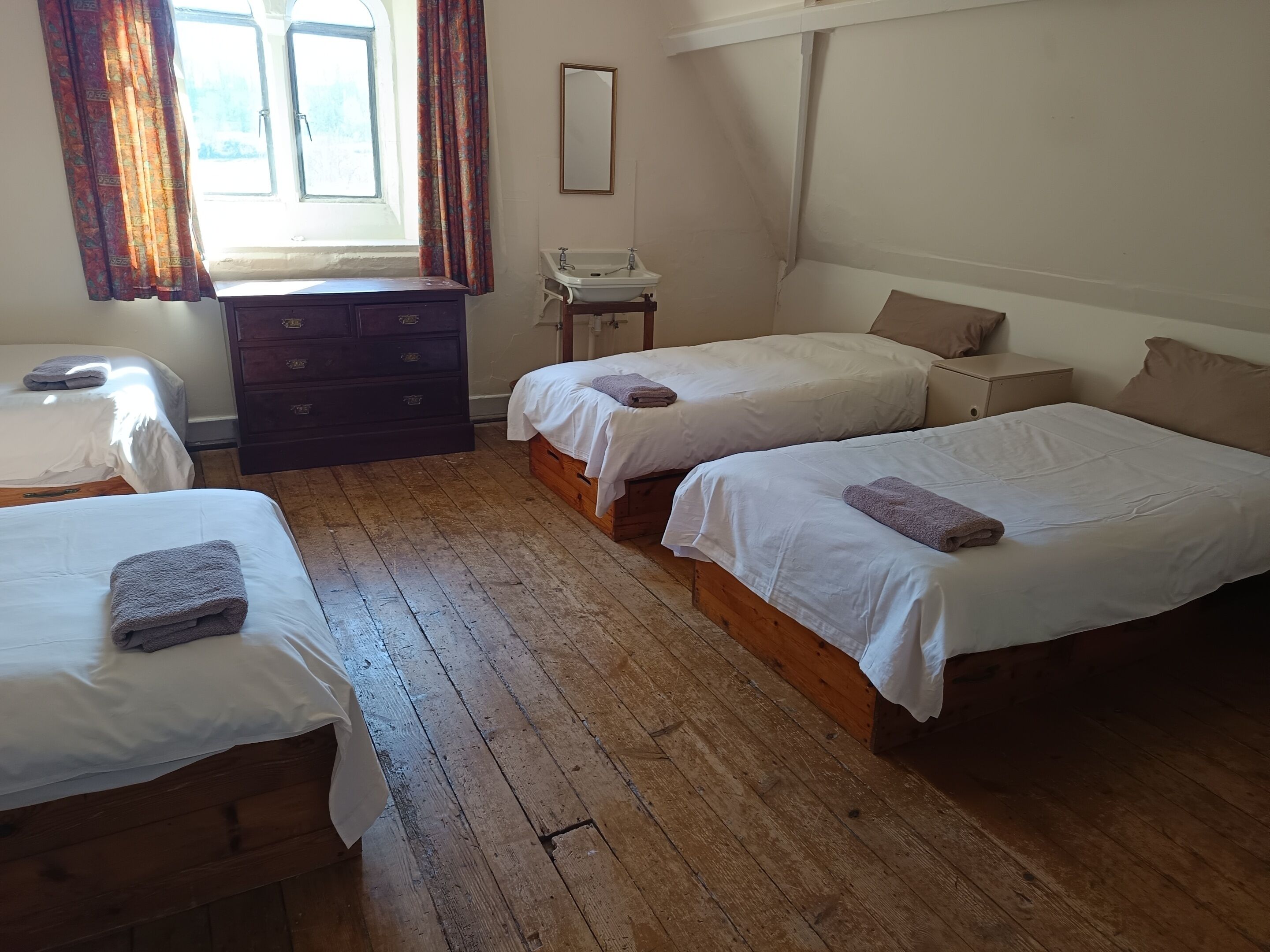 Una habitación con dos camas individuales, un aparador de madera, una mesita con lavamanos y una ventana con cortinas.