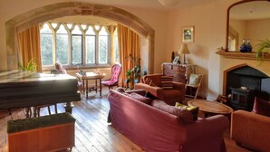 A cozy living room with a grand arched window, a fireplace, a piano, and comfortable seating. - Monkton Wyld Court (Bridport)