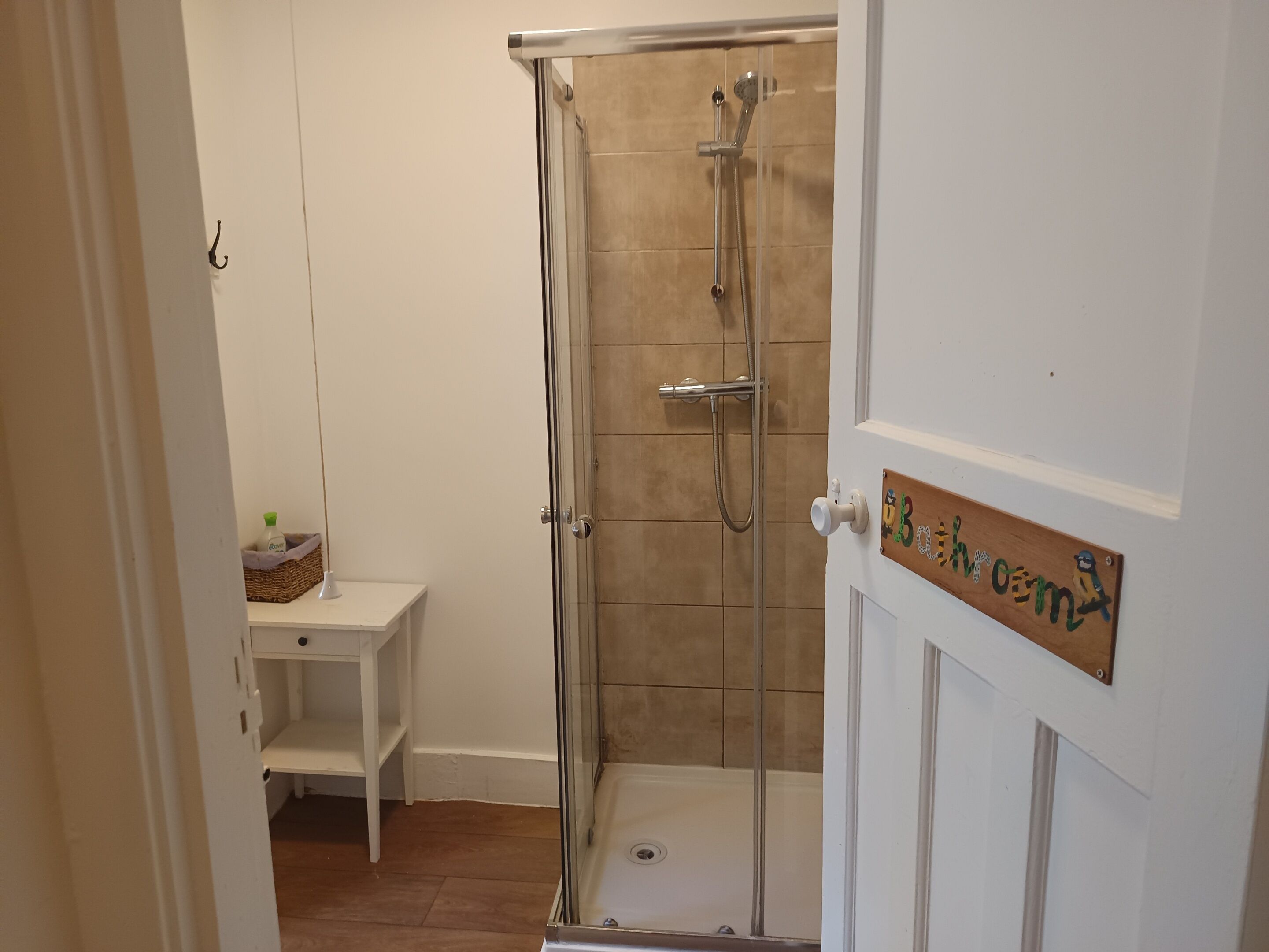 A bathroom with a glass-enclosed shower, a small white shelf with a bottle, and a wooden sign on the door.