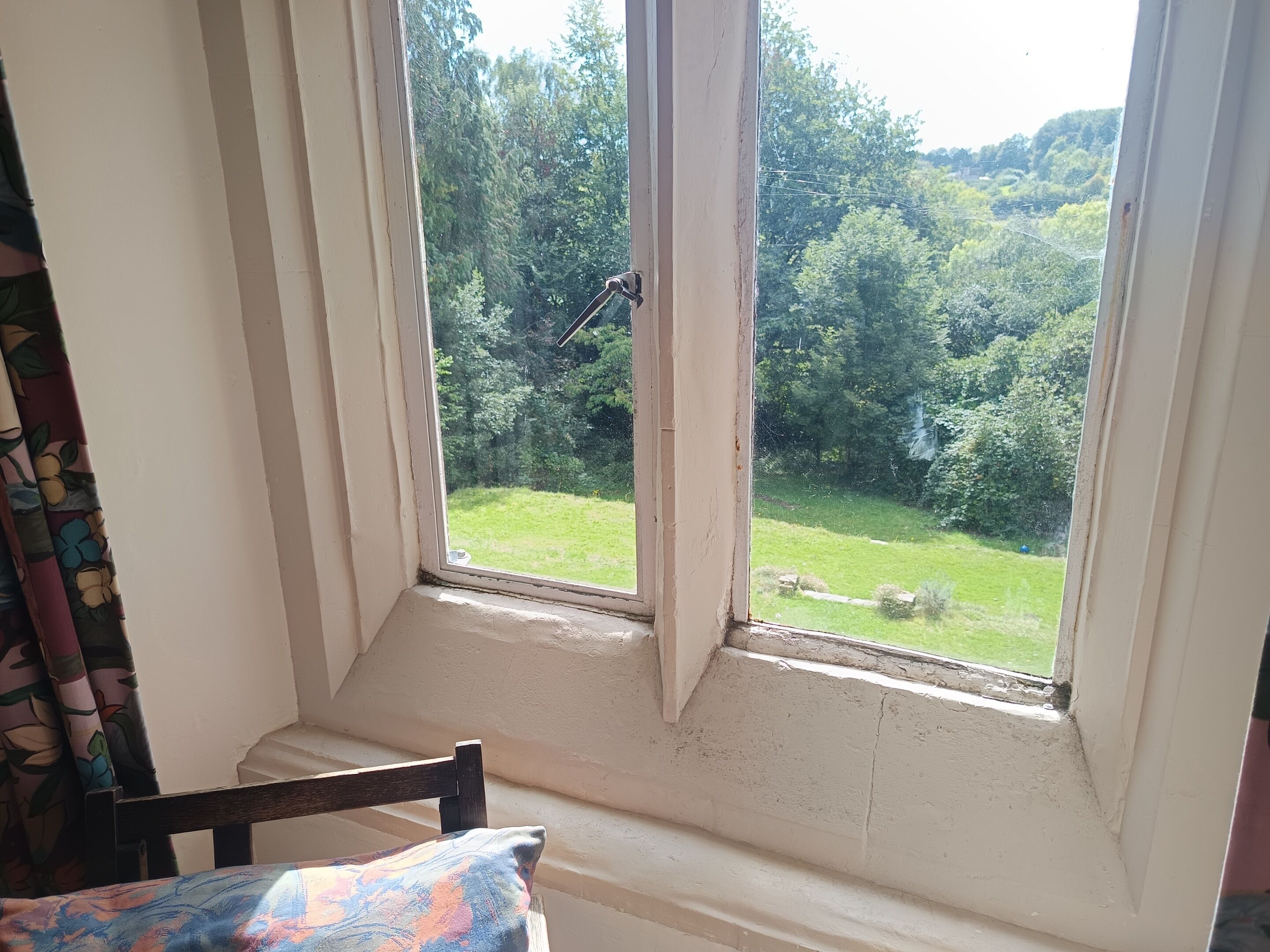 Ein Zimmer mit Fensterblick auf eine grüne Landschaft, ein Stuhl mit gemustertem Kissen und ein teilweise geöffnetes Fenster.