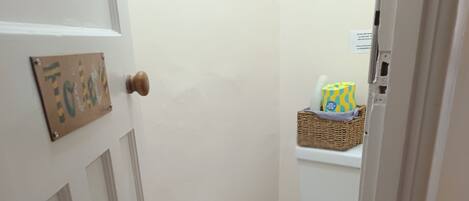 Et lille, rent badeværelse med et hvidt toilet, en skraldespand og en fletkurv på en hylde.