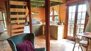 A cozy cabin interior with a wooden dining table, a green armchair, a kitchen area, and a large window. - Monkton Wyld Court (Bridport)
