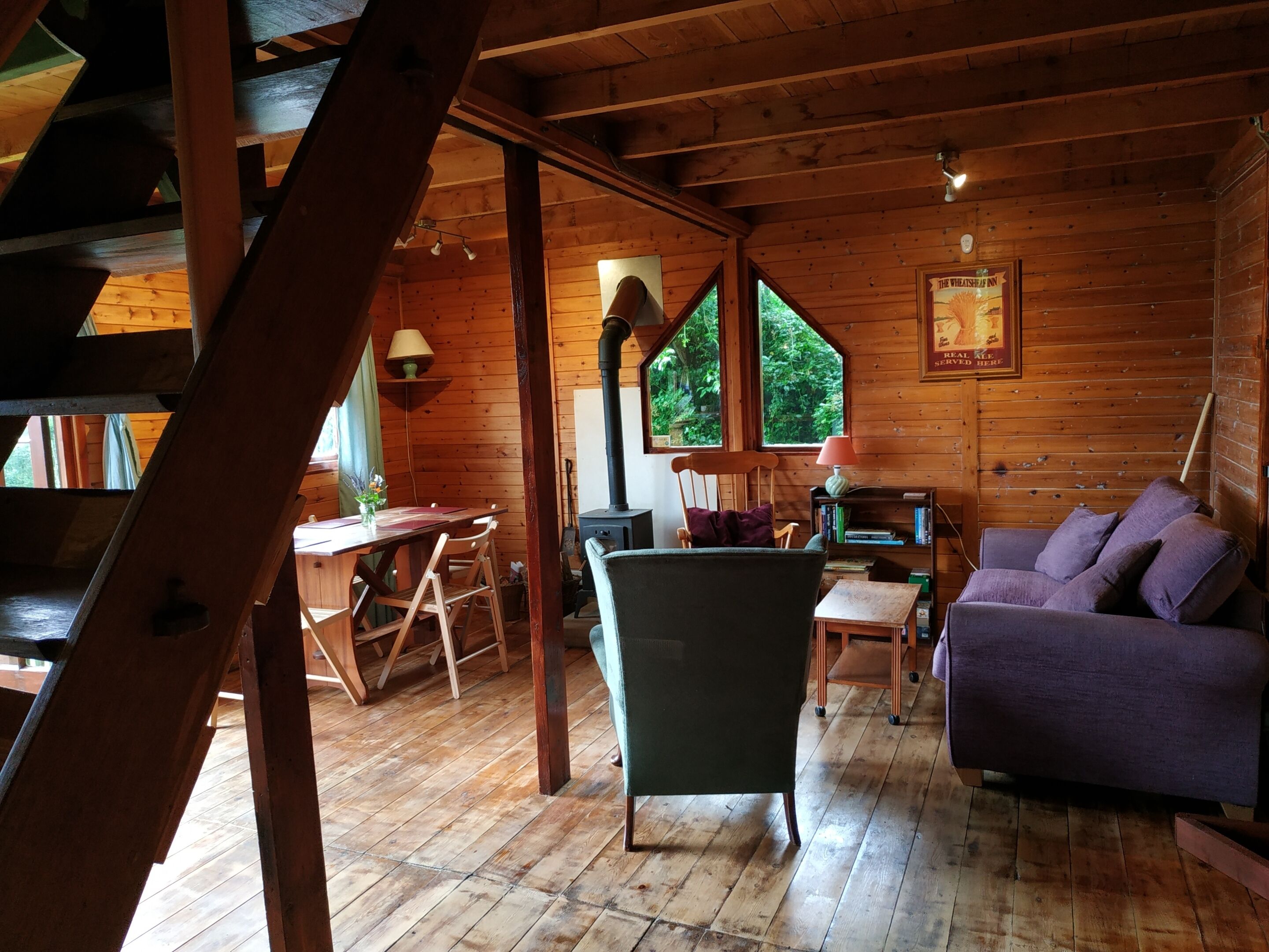A wooden cabin interior with a dining table, chairs, a sofa, a fireplace, and large windows.