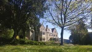 A large stone building with multiple windows and a prominent tower, surrounded by lush greenery and trees. - Monkton Wyld Court (Bridport)