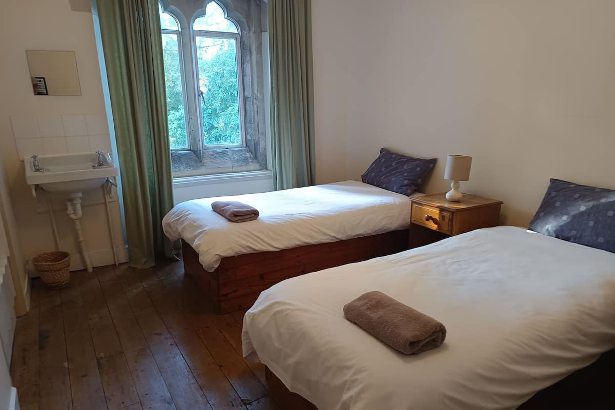 A room with two beds, a window with curtains, a bedside table with a lamp, and a sink.