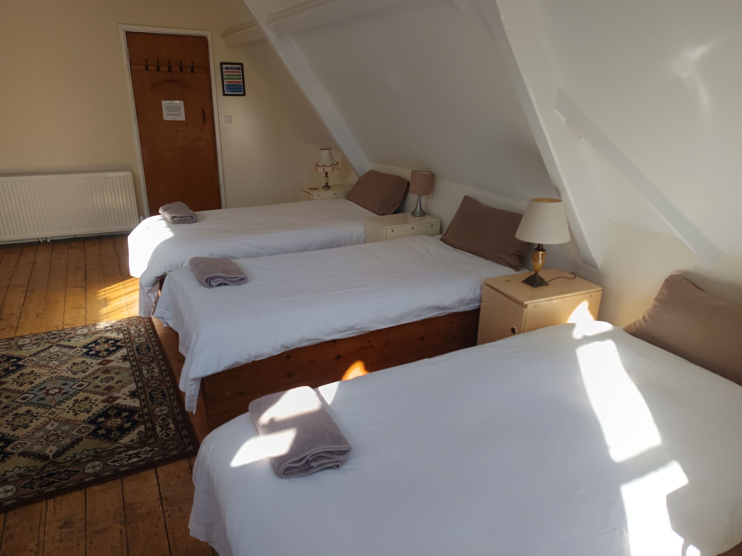 A room with three beds, a wooden floor, a door, a radiator, and a patterned rug.
