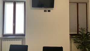 Ein Zimmer mit an der Wand montiertem Fernseher, einem Fenster mit Vorhängen, einem Heizkörper und einem Blumentopf.