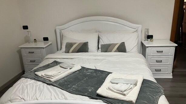 Ein ordentlich bezogenes Bett mit weißen Leinen, zwei Kissen, einer grauen und weißen Tagesdecke und einem gefalteten Handtuch. Zwei Nachttische mit Schubladen sind vorhanden, jeder mit einer kleinen Lampe und einem dekorativen Gegenstand.