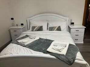 Un lit bien fait, avec du linge de lit blanc, deux oreillers, un plaid gris et blanc et une serviette pliée. Il y a deux tables de chevet avec des tiroirs, chacune étant surmontée d'une petite lampe et d'un objet décoratif.