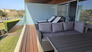 Un área de estar moderna al aire libre con un sofá y sillones, una terraza de madera y una pared de vidrio que ofrece vistas del paisaje.