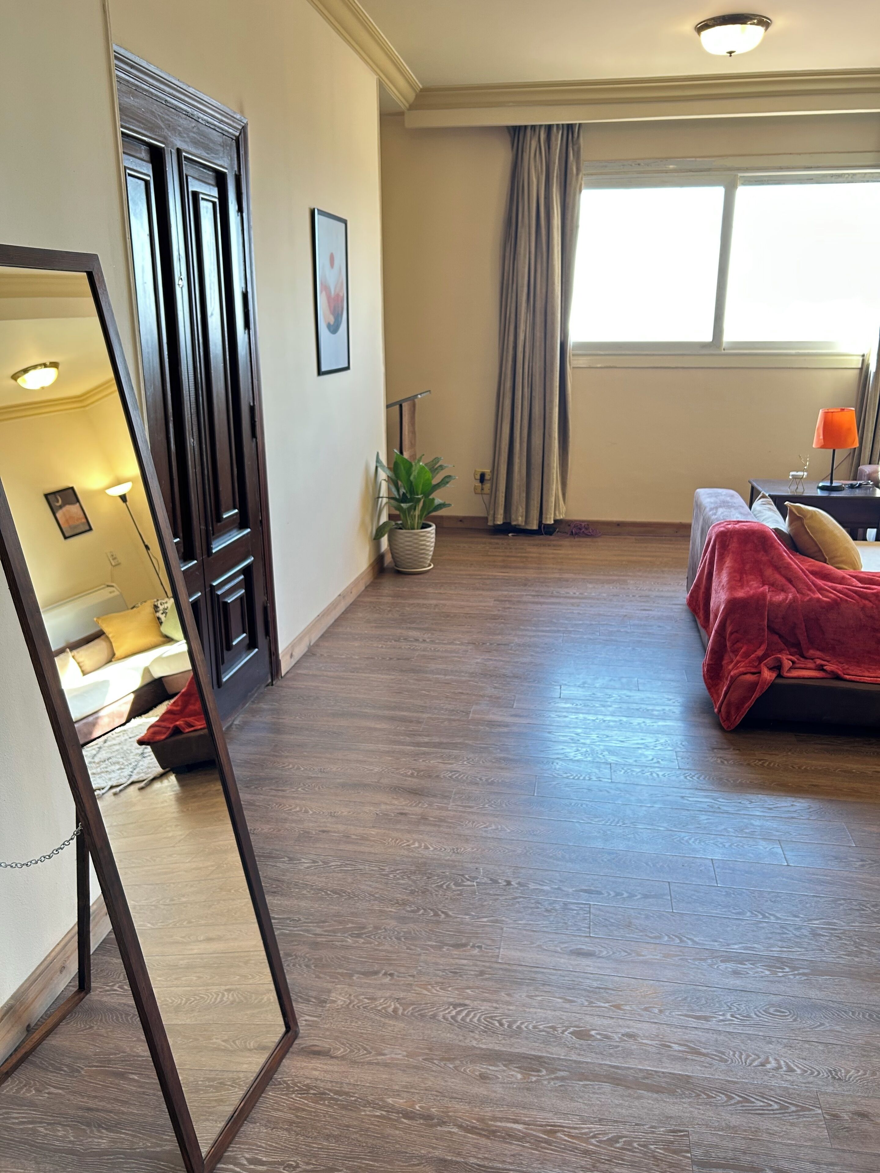 Une pièce avec un plancher en bois, un lit recouvert d’une couverture rouge, une plante en pot, une fenêtre avec des rideaux et un miroir qui reflète une partie de la pièce.