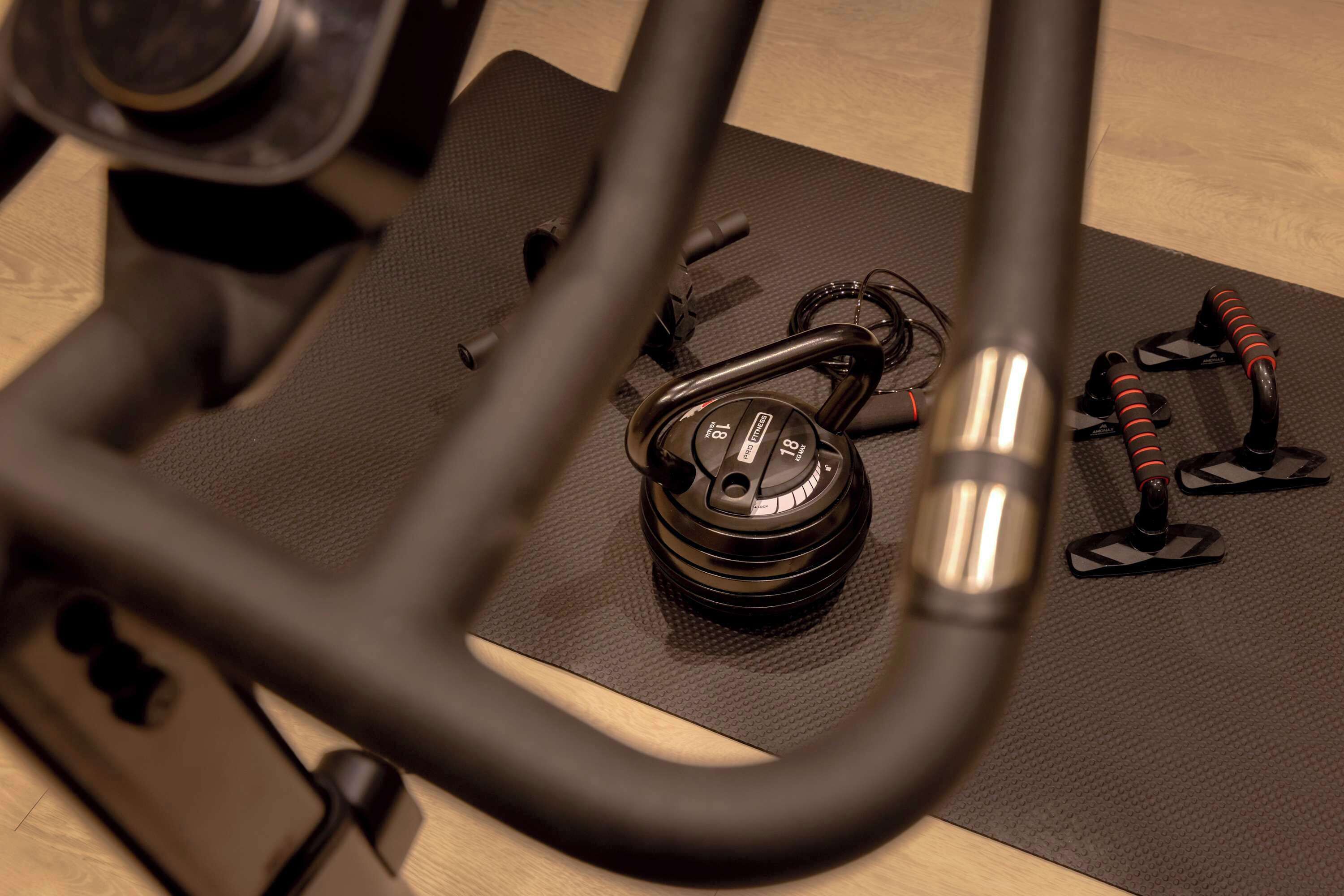 A black exercise bike with a digital display, coiled cables, and a mat on a wooden floor.