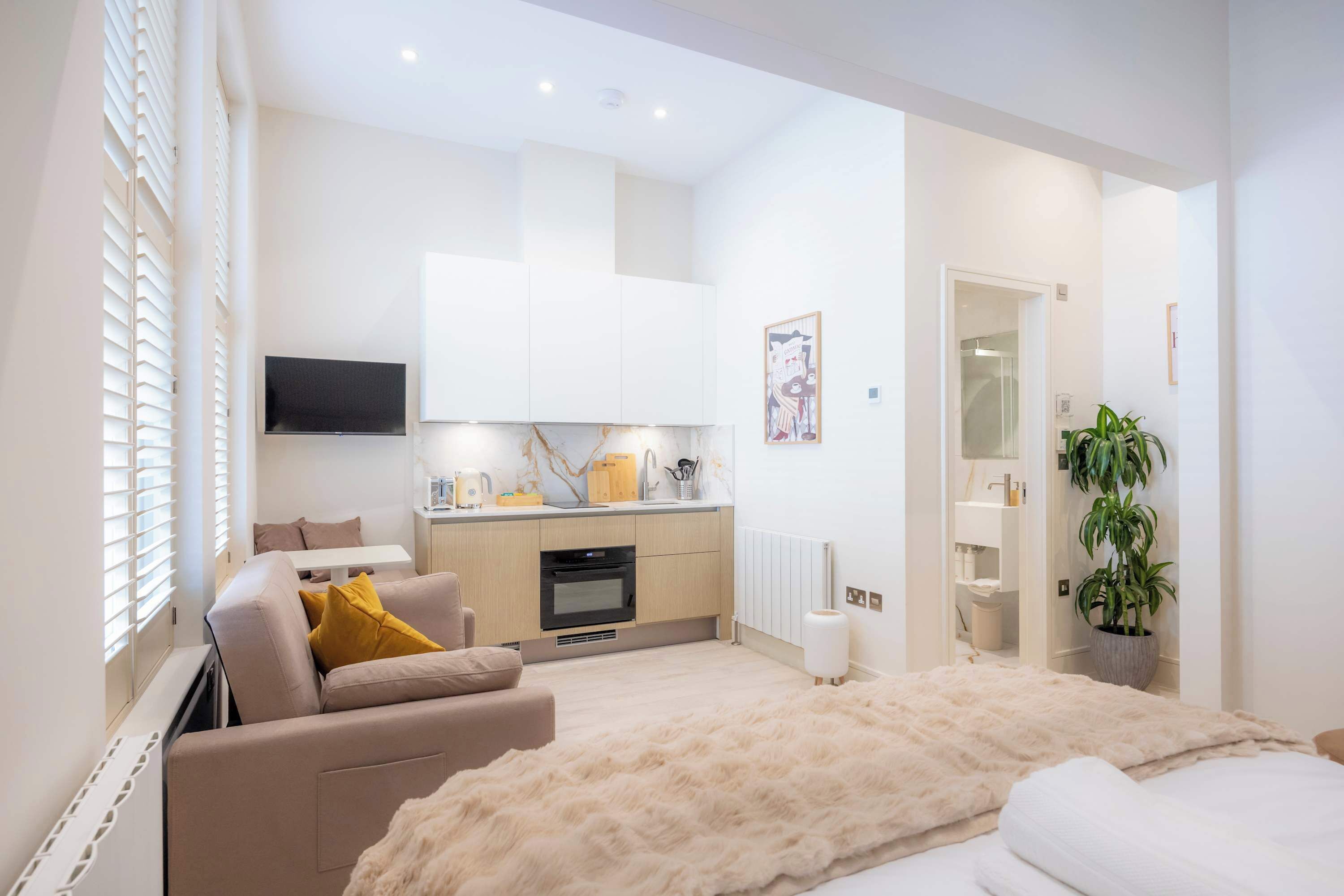 A modern hotel room with a sofa, a large bed, a kitchen area, and a TV.