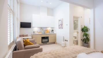 A modern hotel room with a sofa, a large bed, a kitchen area, and a TV.