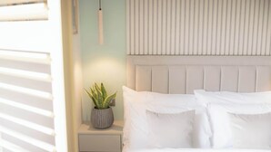 Un dormitorio con cama, almohadas blancas, mesita de noche con planta y cabecera.