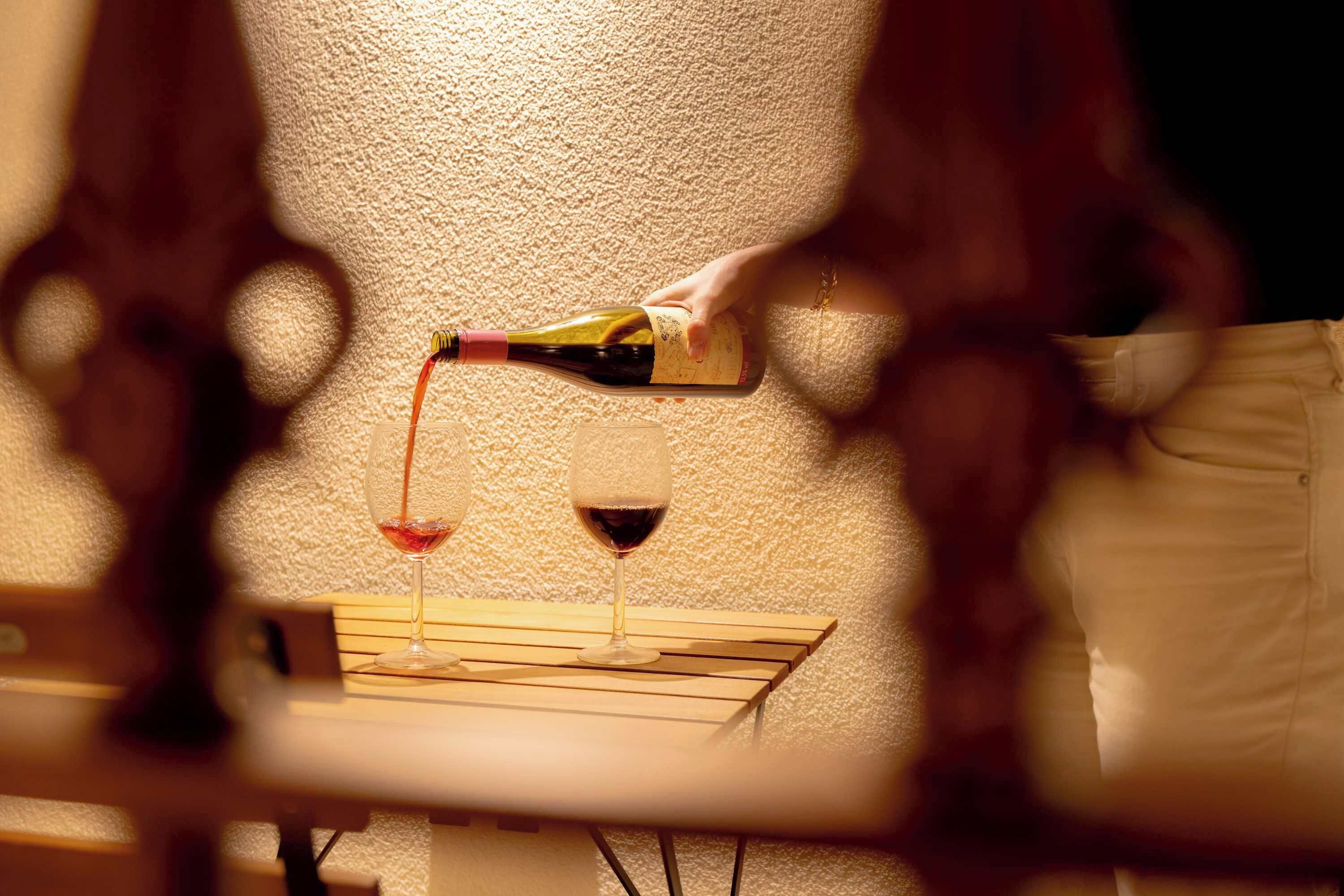 A bottle pouring wine into a glass on a wooden table against a textured wall.