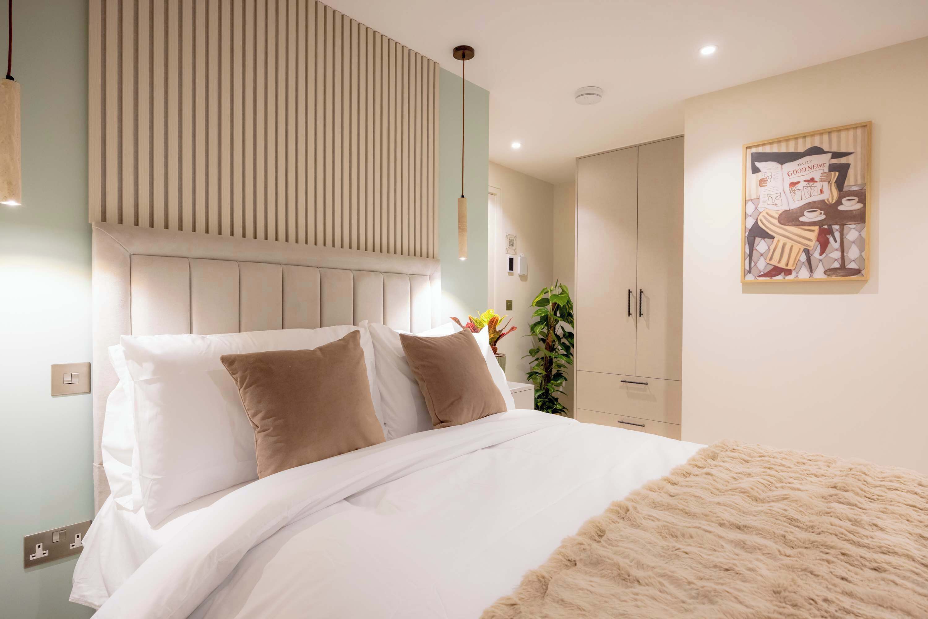 A neatly made bed with white and beige bedding, a wall-mounted light fixture, a framed picture, and a plant.