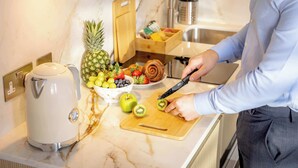 Une cuisine équipée d’une machine à café, d’une planche à découper, d’une personne qui épluche un kiwi et d’un saladier rempli de fruits.