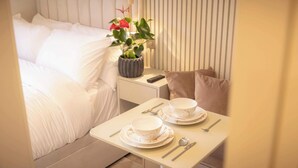 Un lit bien fait, avec une literie blanche, une table de chevet avec une télécommande, une plante en pot et une table préparée pour le petit-déjeuner avec des assiettes, des bols et des couverts.