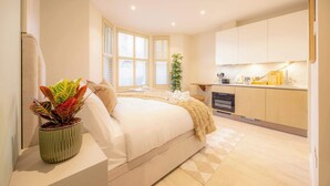 Quarto com cama, planta na mesinha de cabeceira e vista para a cozinha.