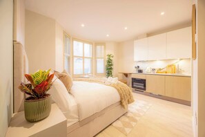 Un dormitorio con cama, planta en la mesita de noche y vista a la cocina.