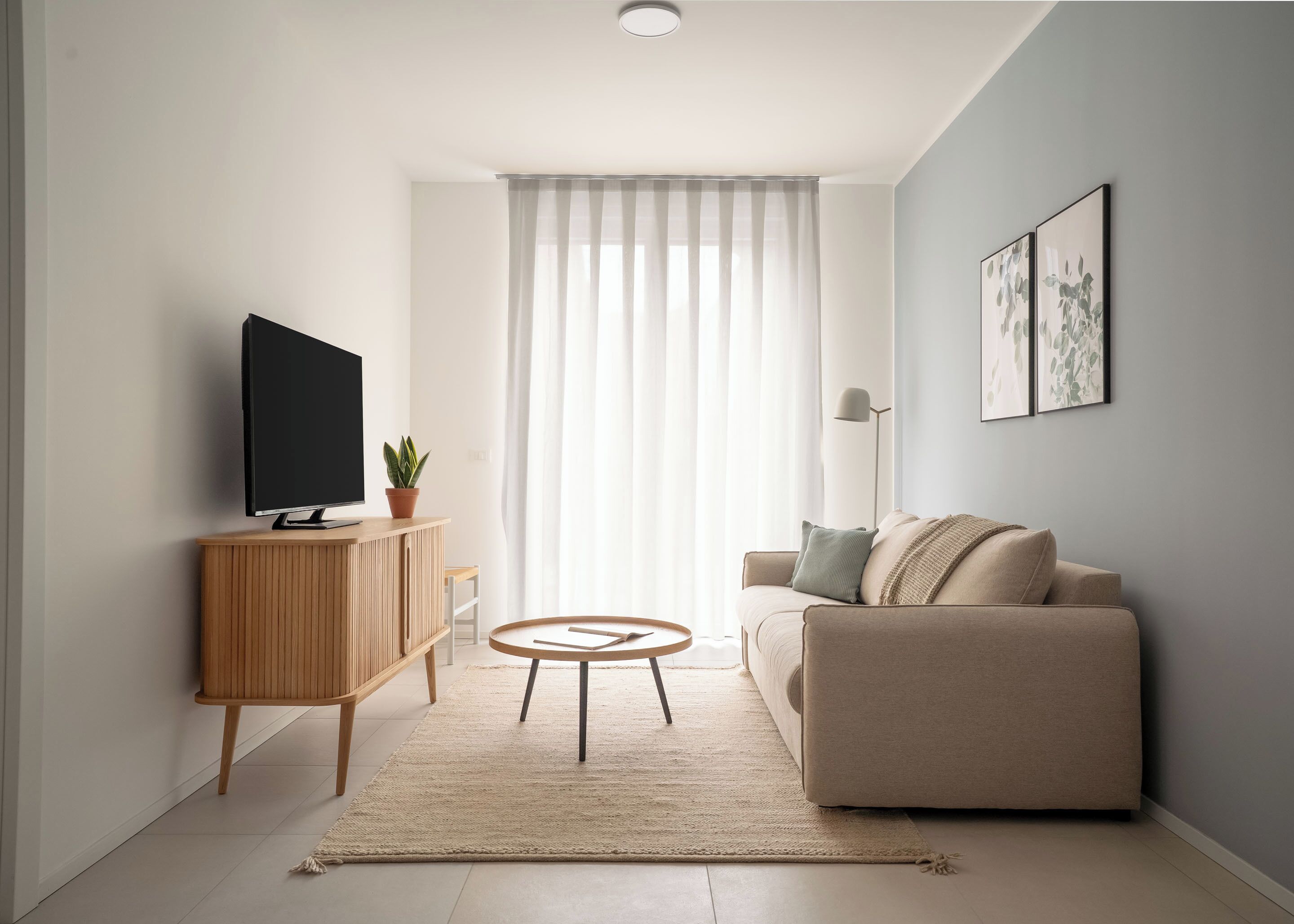 Una sala de estar moderna con un televisor de pantalla plana, un mueble de madera, una mesa de centro redonda, un sofá beige y un ventanal con cortinas traslúcidas.