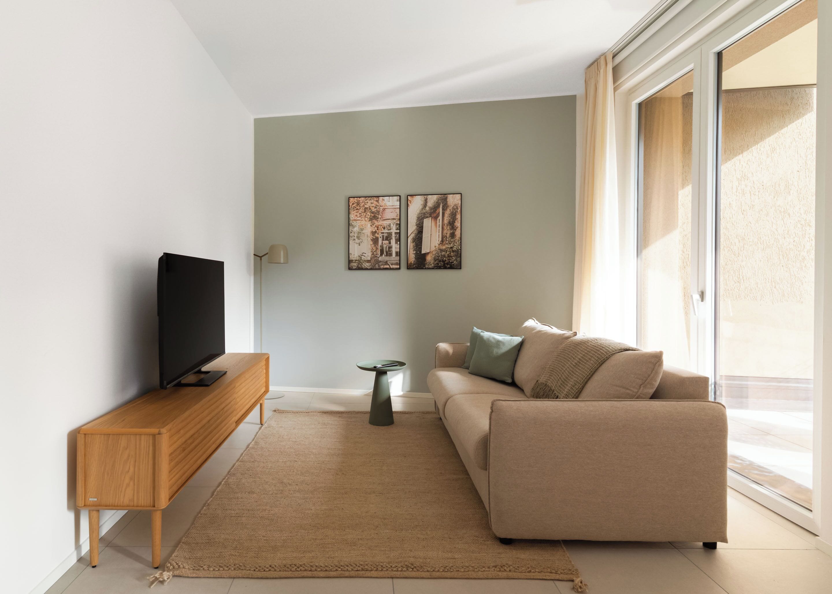 A modern living room with a flat-screen TV on a wooden console, a beige sofa with cushions, a small side table, and two framed artworks on the wall.