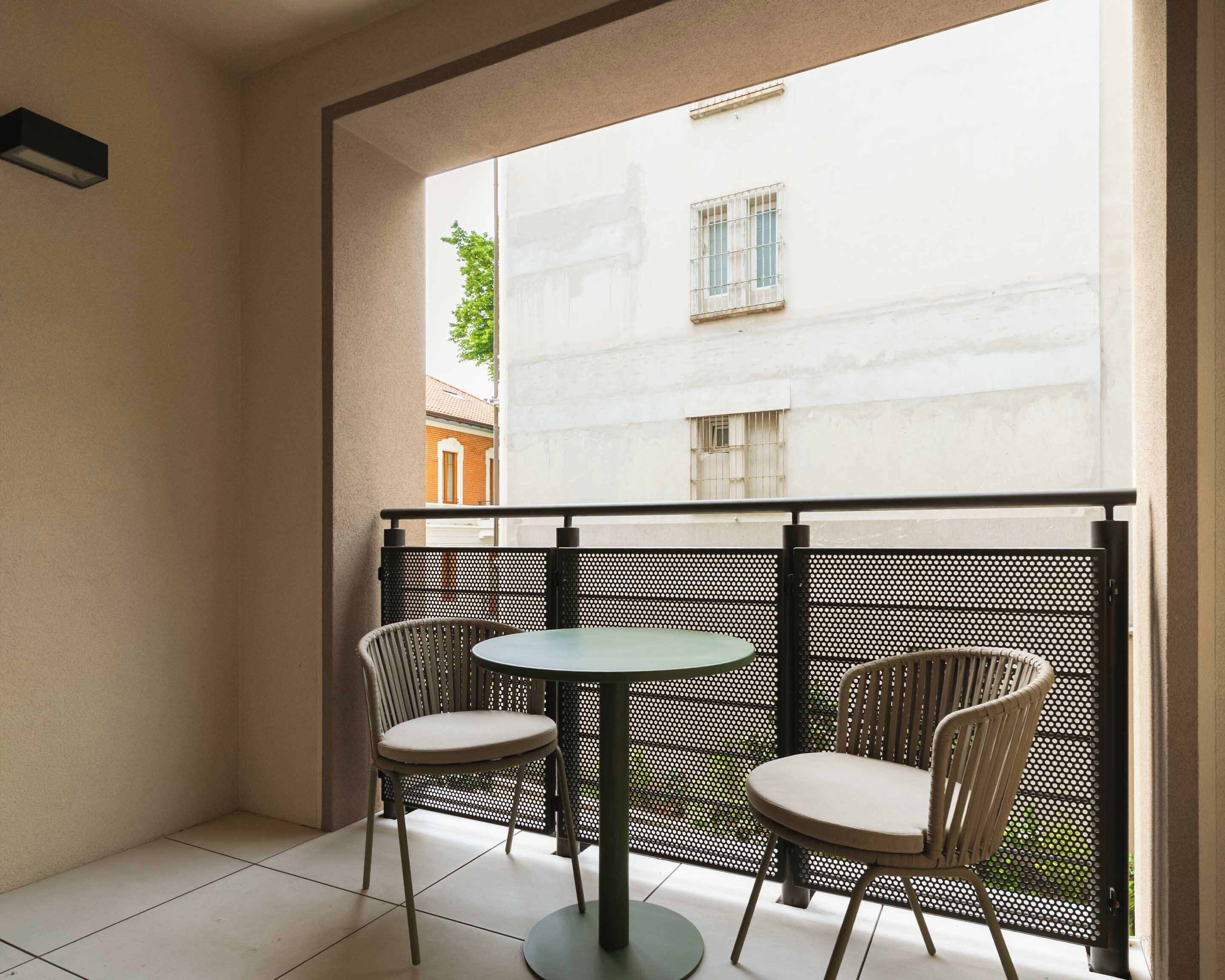 A balcony with two chairs and a table, a metal fence, and a view of a building with barred windows.