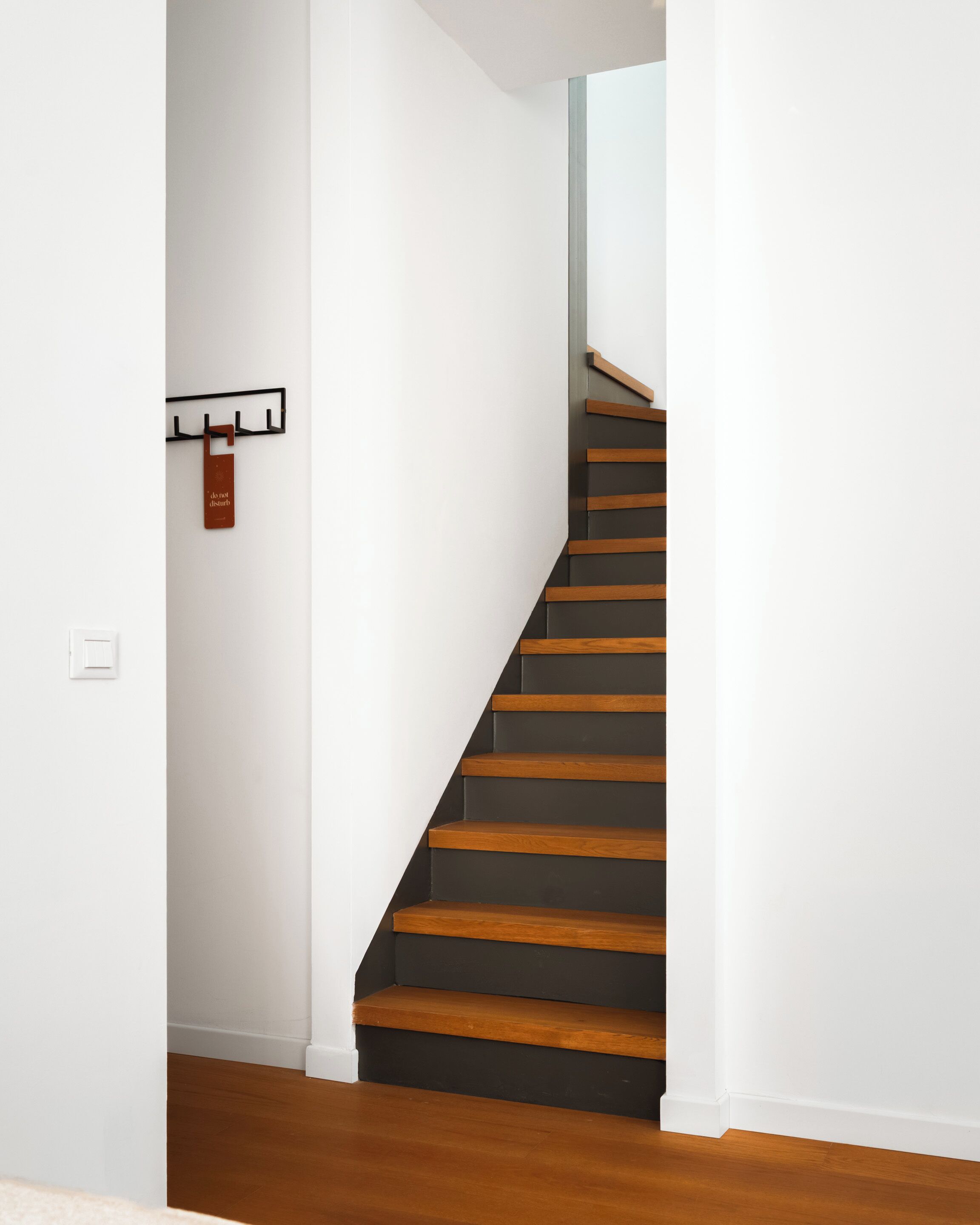 A staircase with wooden steps and a metal handrail, leading to a room with white walls and a wooden floor.