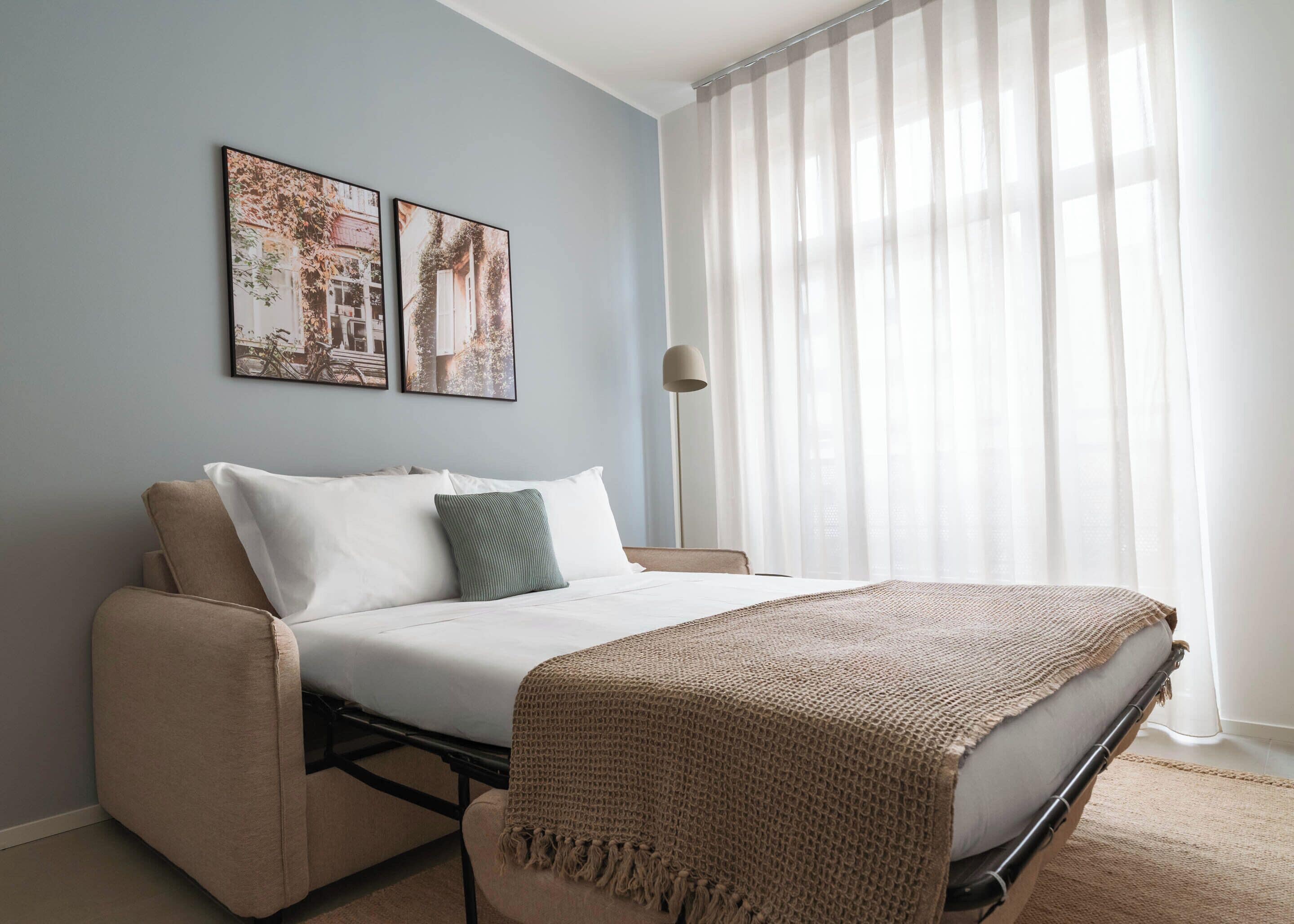 A bedroom with a bed, a sofa, a floor lamp, and two framed pictures on the wall.