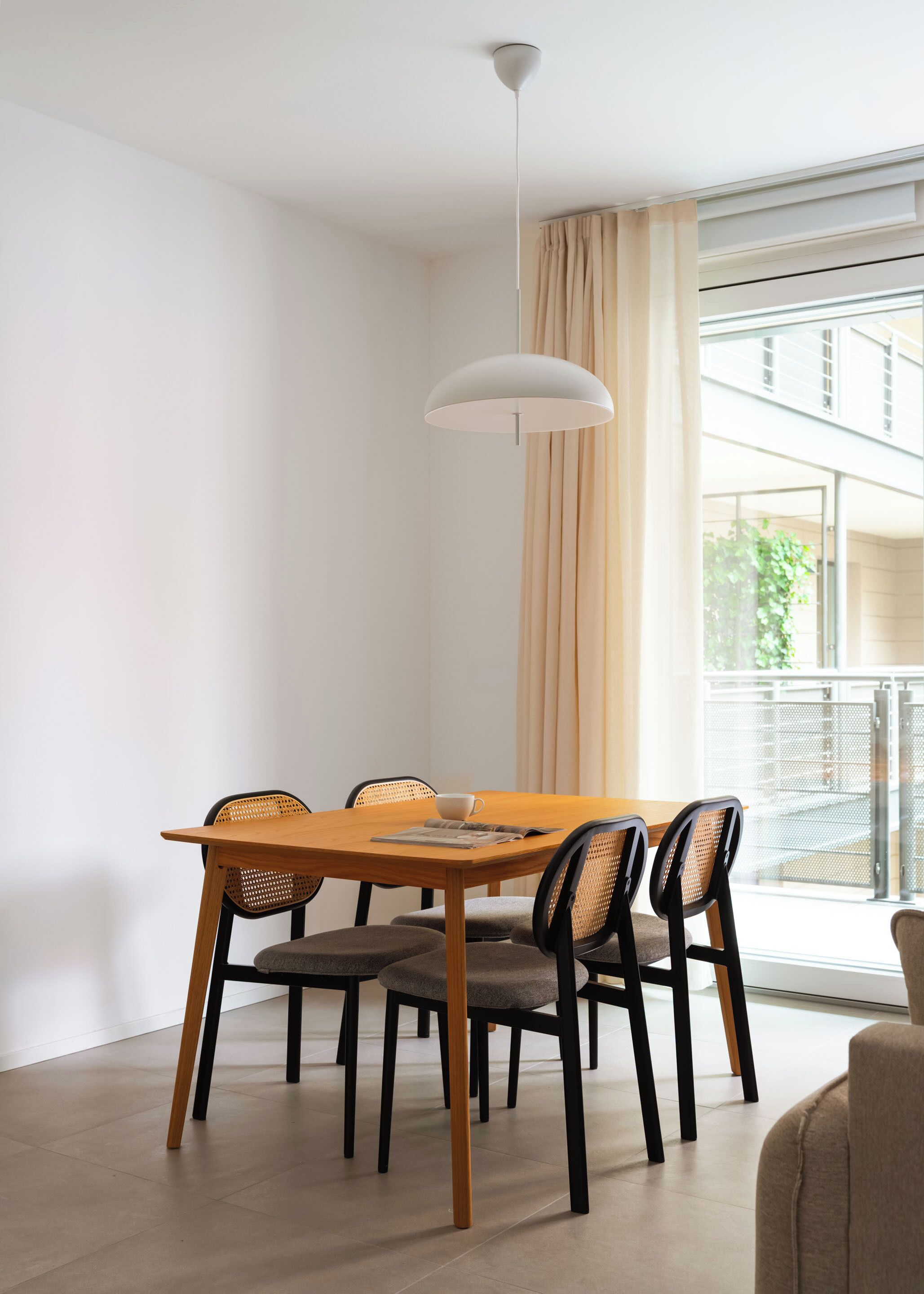 Un comedor con una mesa de madera, cuatro sillas, una lámpara colgante blanca y un ventanal con vistas a edificios.