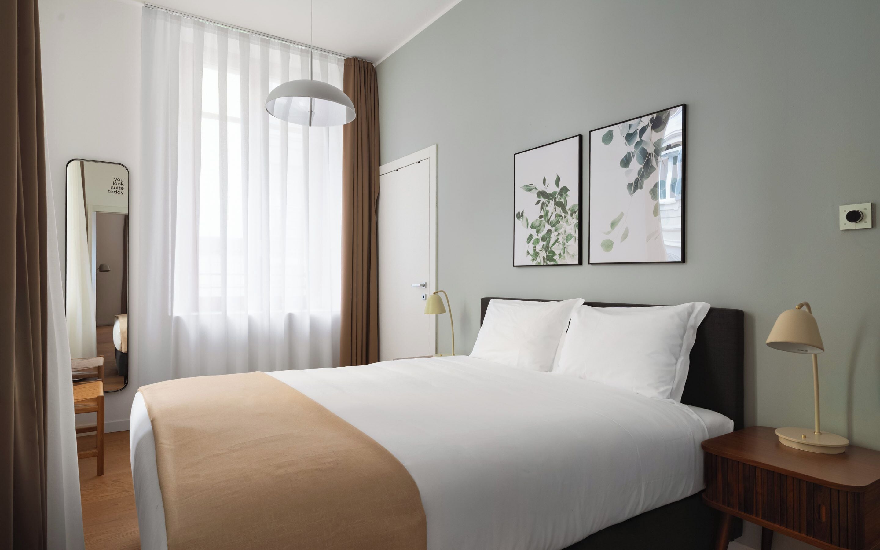 A hotel room with a bed, bedside lamps, a mirror, and two framed pictures on the wall.