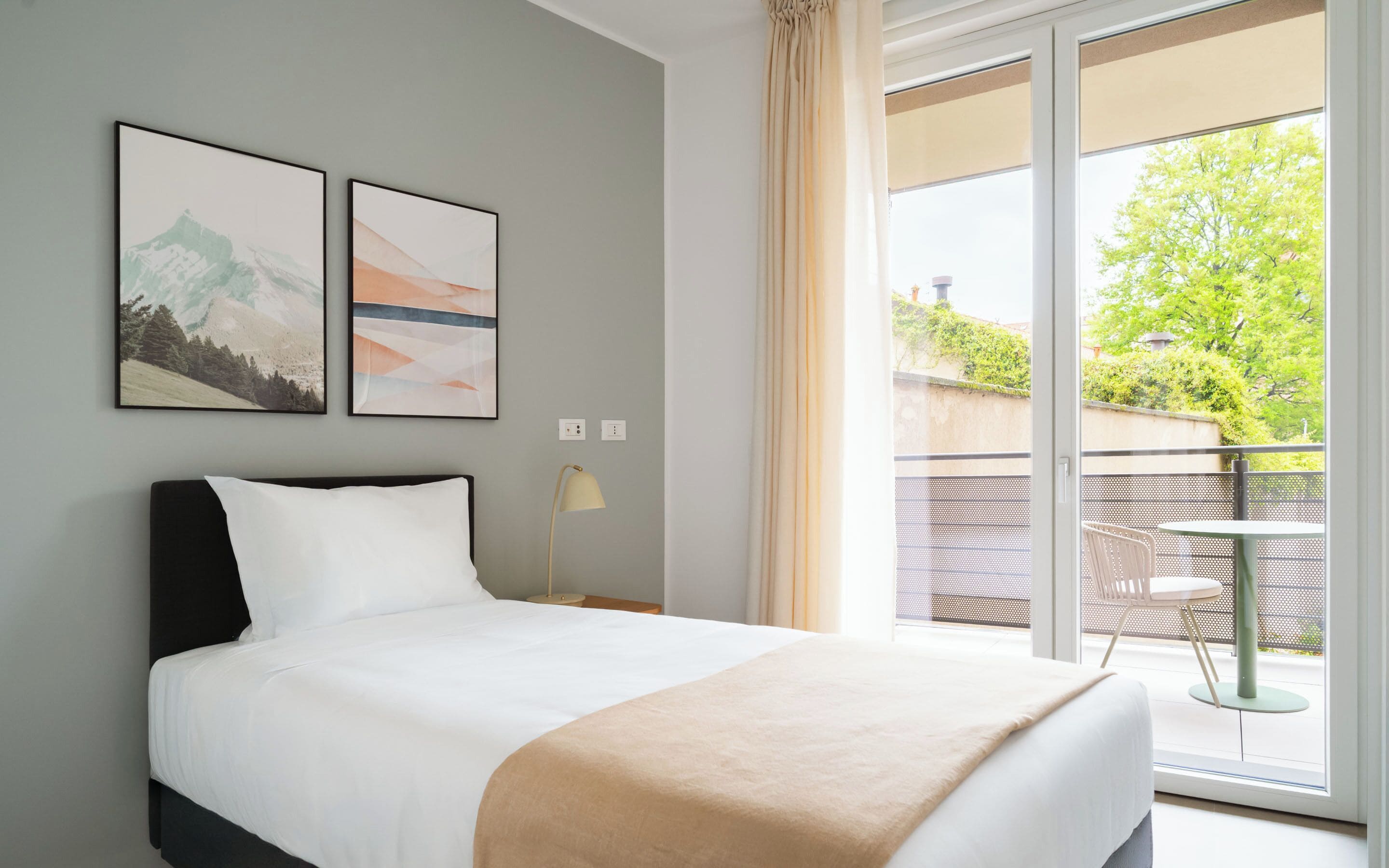 A hotel room with a bed, a nightstand, a lamp, two framed pictures, and a balcony with a view of greenery.