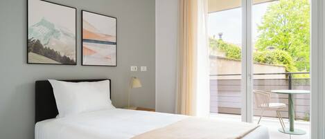 A hotel room with a bed, a nightstand, a lamp, two framed pictures, and a balcony with a view of greenery.