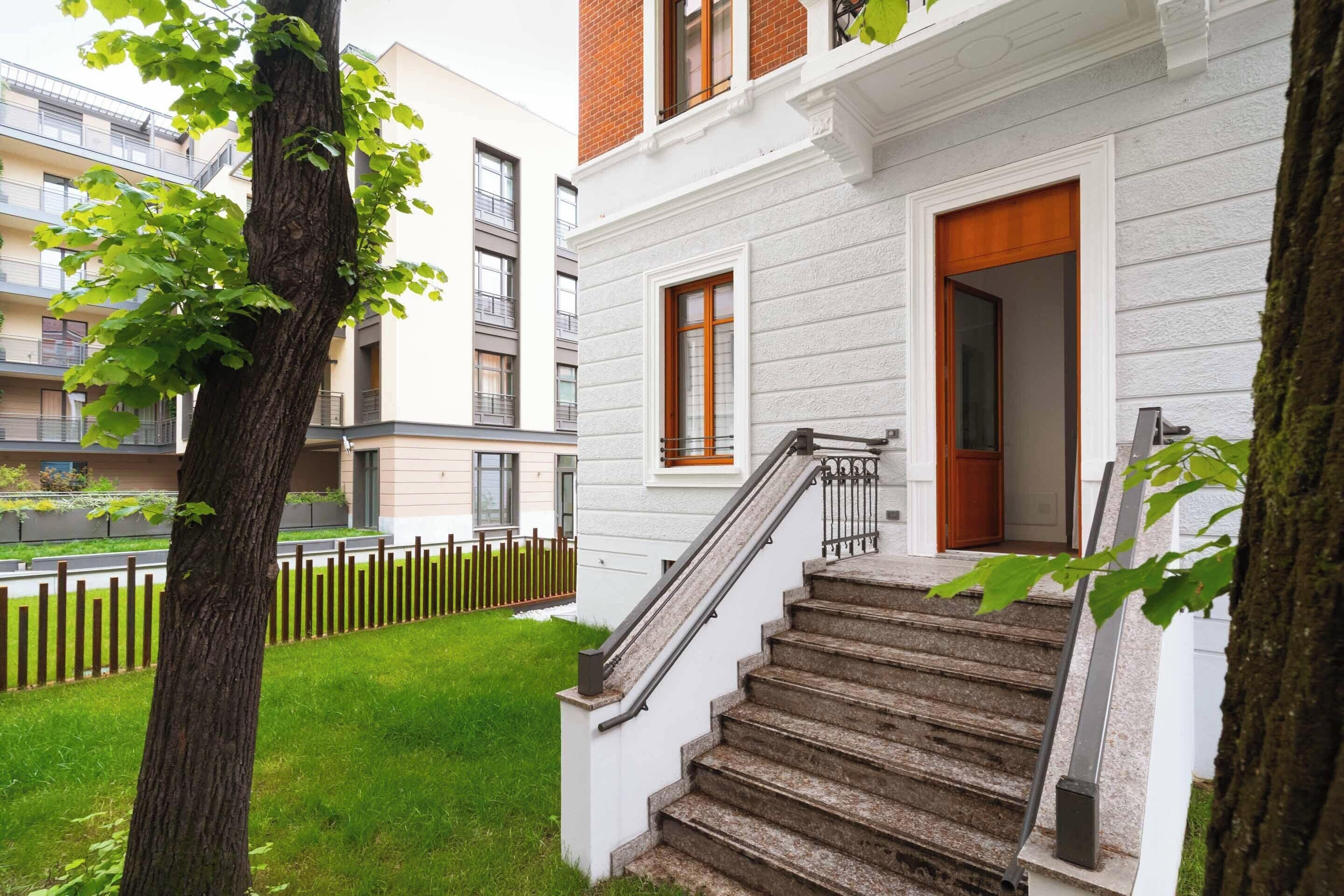 A residential building with a staircase leading to a wooden door, a small window, and a tree in the foreground.