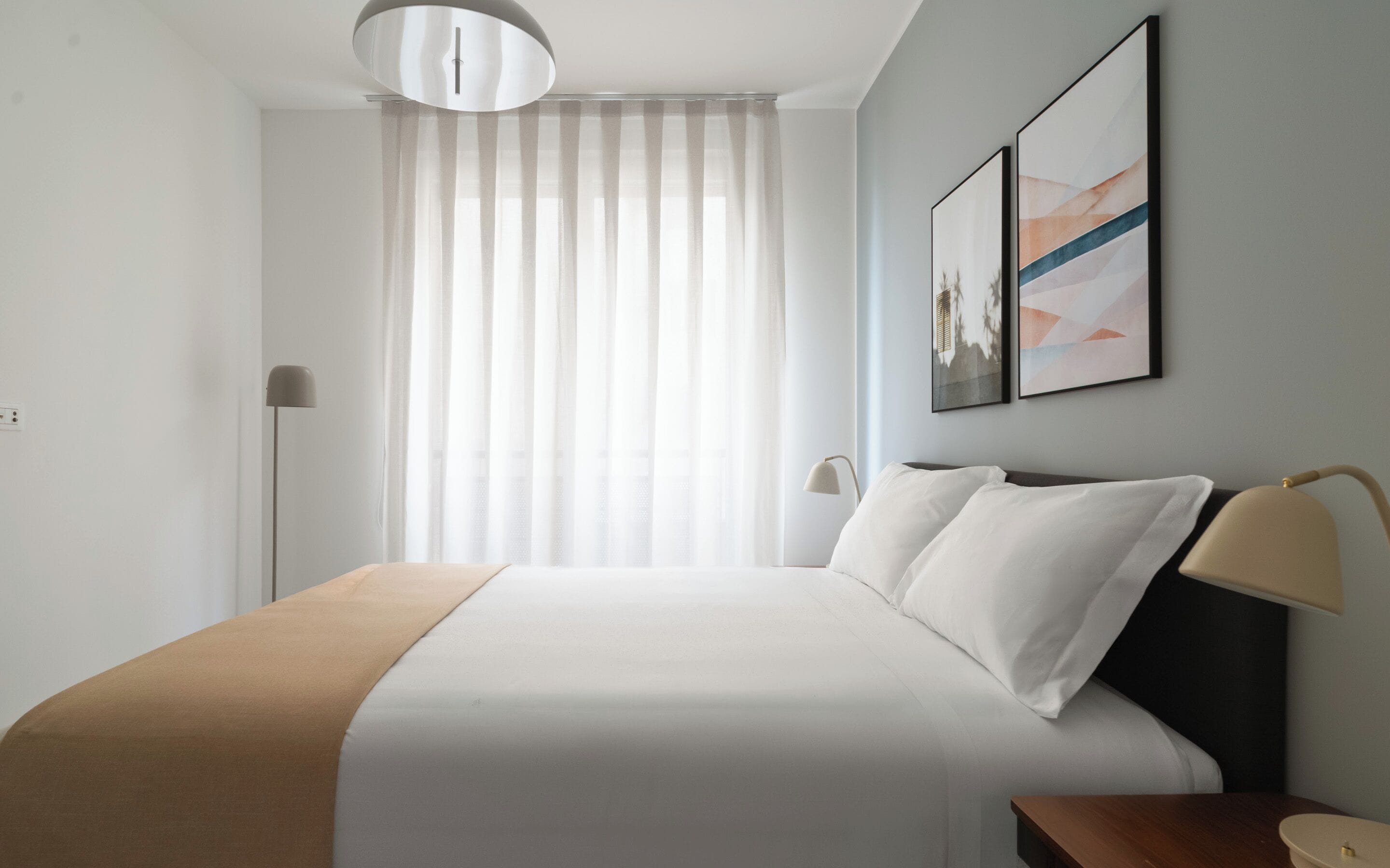 Una habitación de hotel con una cama, una mesita de noche, una lámpara, una ventana con cortinas translúcidas y dos obras de arte colgadas en la pared.