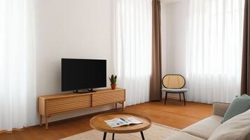 Un salon moderne avec un téléviseur à écran plat posé sur une console en bois, une table basse ronde avec un magazine, un canapé gris, un fauteuil en osier et des rideaux légers.