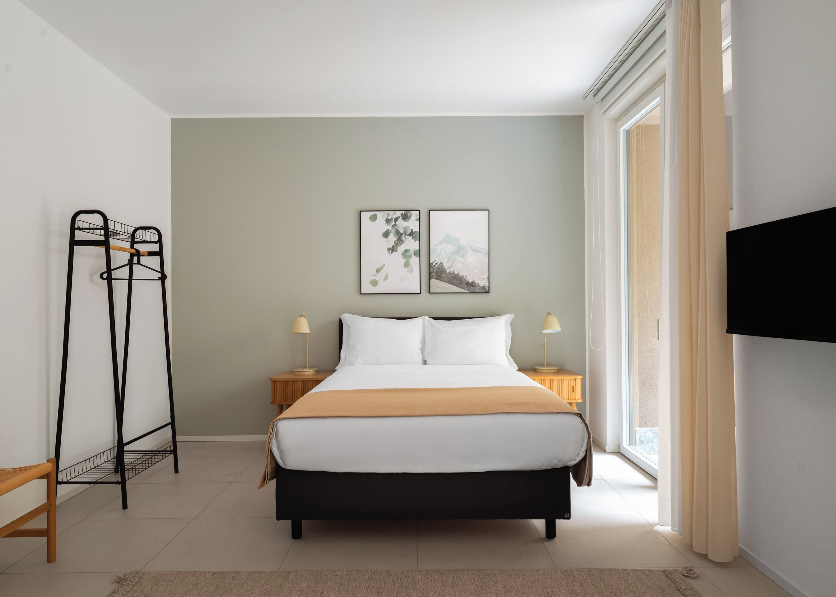 A bedroom with a bed, two bedside tables, a black metal drying rack, and two framed pictures on the wall.