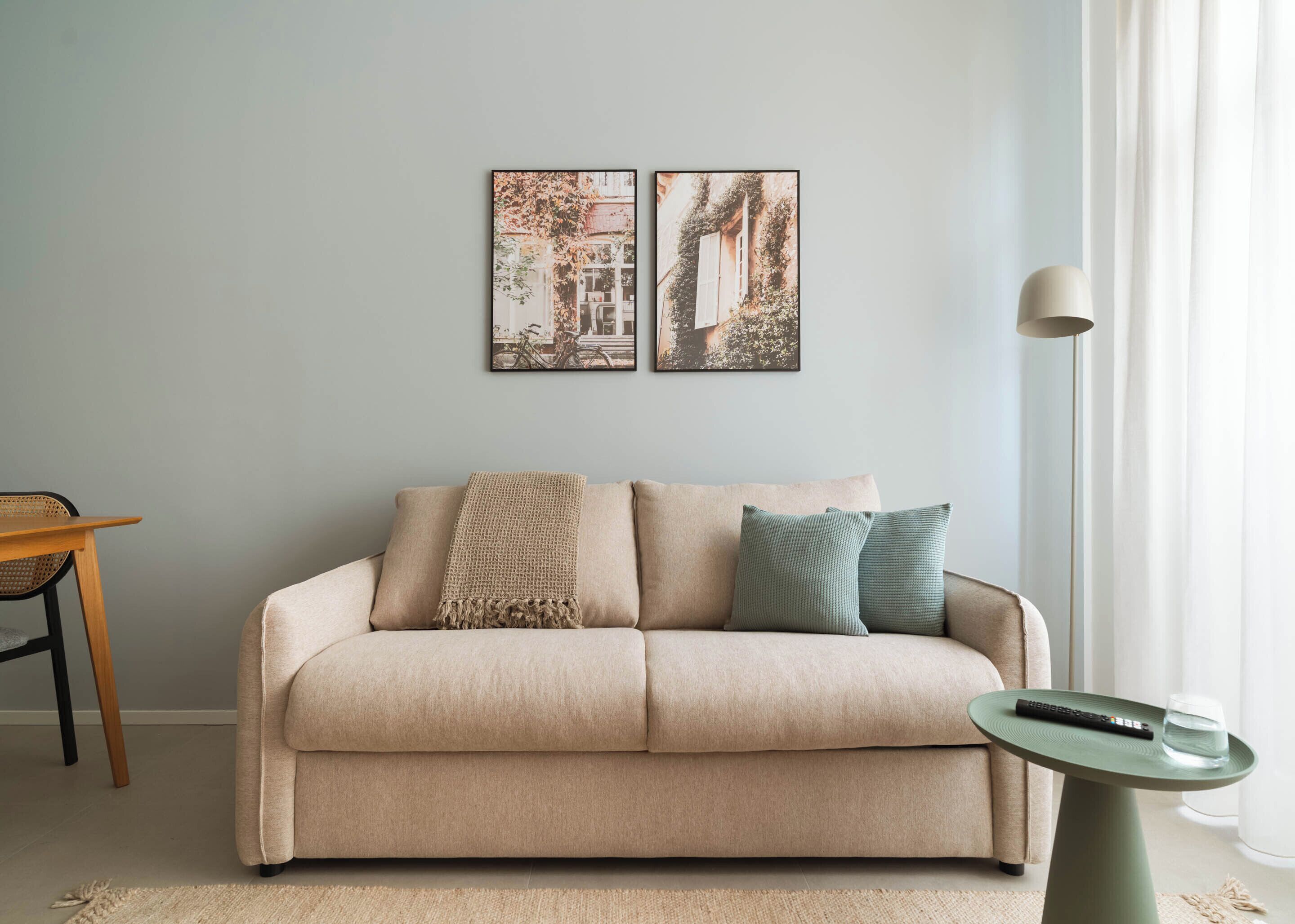 Un salón con un sofá color beige, una mesita redonda con un control remoto y un vaso, y dos cuadros en la pared.