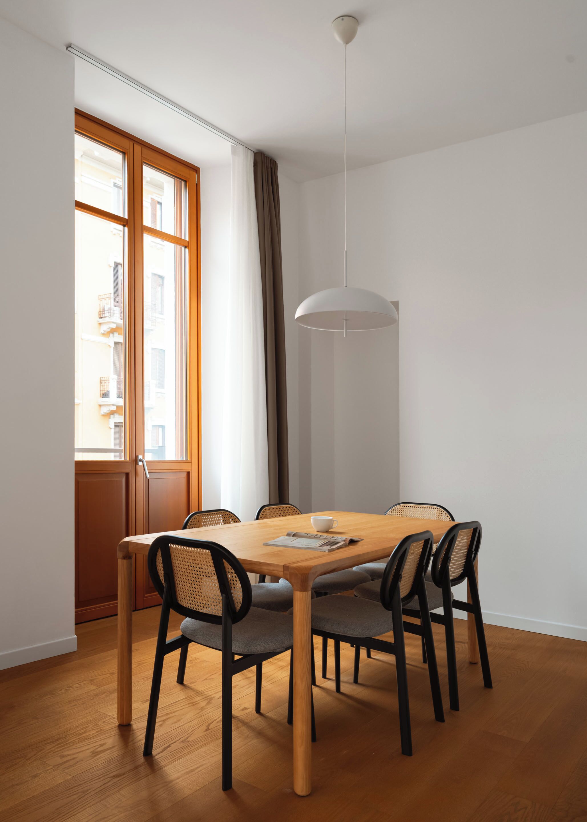 A dining area with a wooden table, six chairs, a hanging light fixture, and a window with curtains.