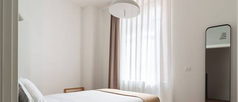 A bedroom with a bed, a bedside table, a mirror, and a window with curtains.