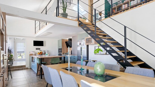 Une cuisine moderne dotée d’un îlot central, d’un coin repas avec une table en verre et d’un escalier avec une rampe en métal noir.