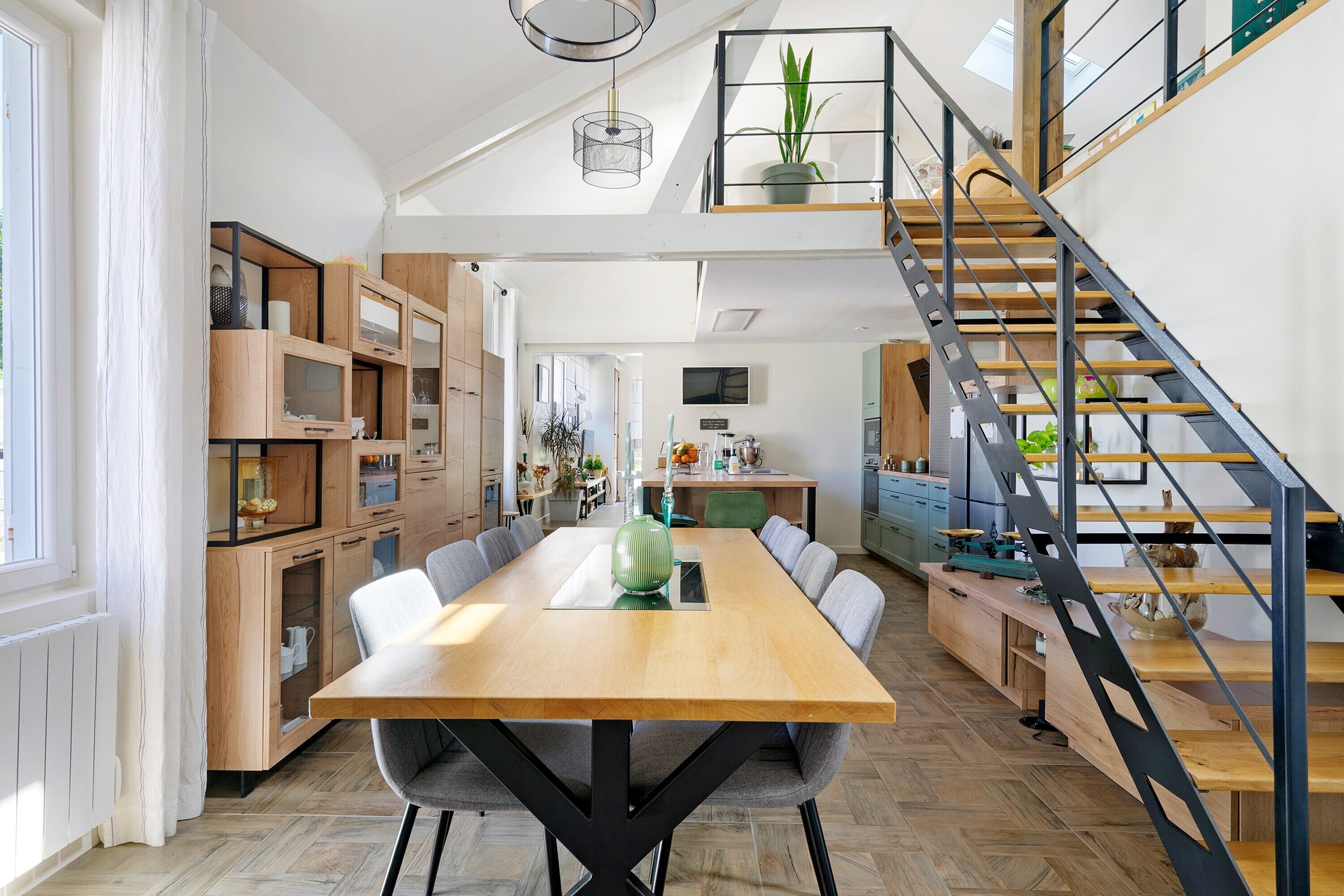 A spacious dining area with a wooden table and chairs, a staircase with a metal railing, and a kitchen in the background.