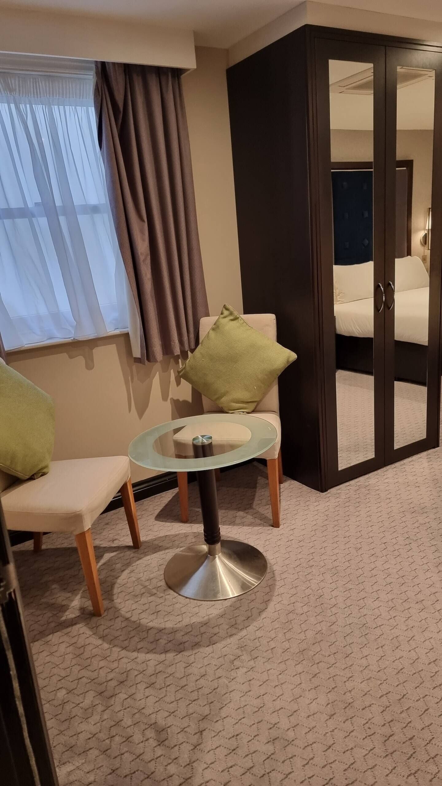 Een hotelkamer met een bed, een glazen tafel, een stoel, een raam met gordijnen en een spiegel.