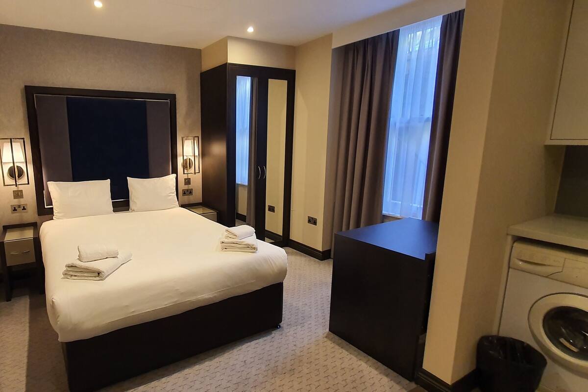 Quarto de hotel com uma cama grande, luminárias de cabeceira, um guarda-roupa, máquina de lavar e uma janela com cortinas.