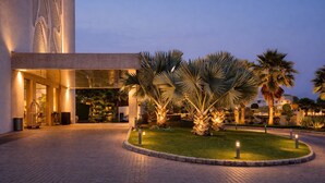 Entrada de hotel con camino de entrada pavimentado, palmeras y fachada de edificio bien iluminada.