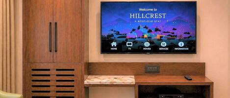 Habitación de hotel con televisión mostrando 'Welcome to Hillcrest Boutique Stay', un aparador de madera, una mesita con un libro y una planta en maceta.
