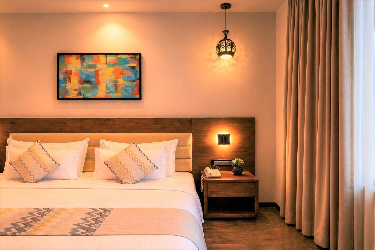 Une chambre d’hôtel avec un grand lit, une table de chevet avec un téléphone et une lampe, un tableau au mur et une fenêtre avec des rideaux.
